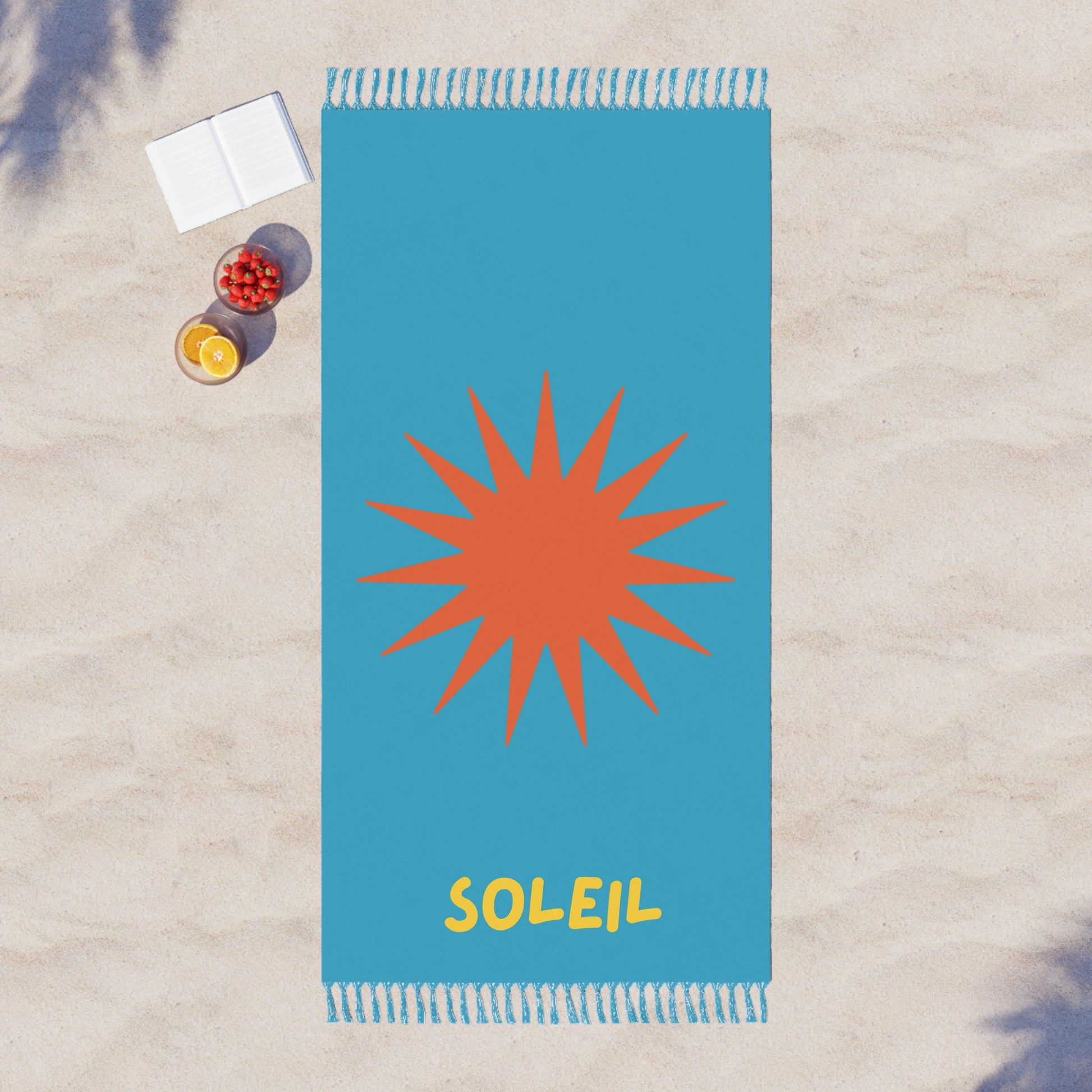 Blue beach towel with orange sun design and 'SOLEIL' text on a sandy background