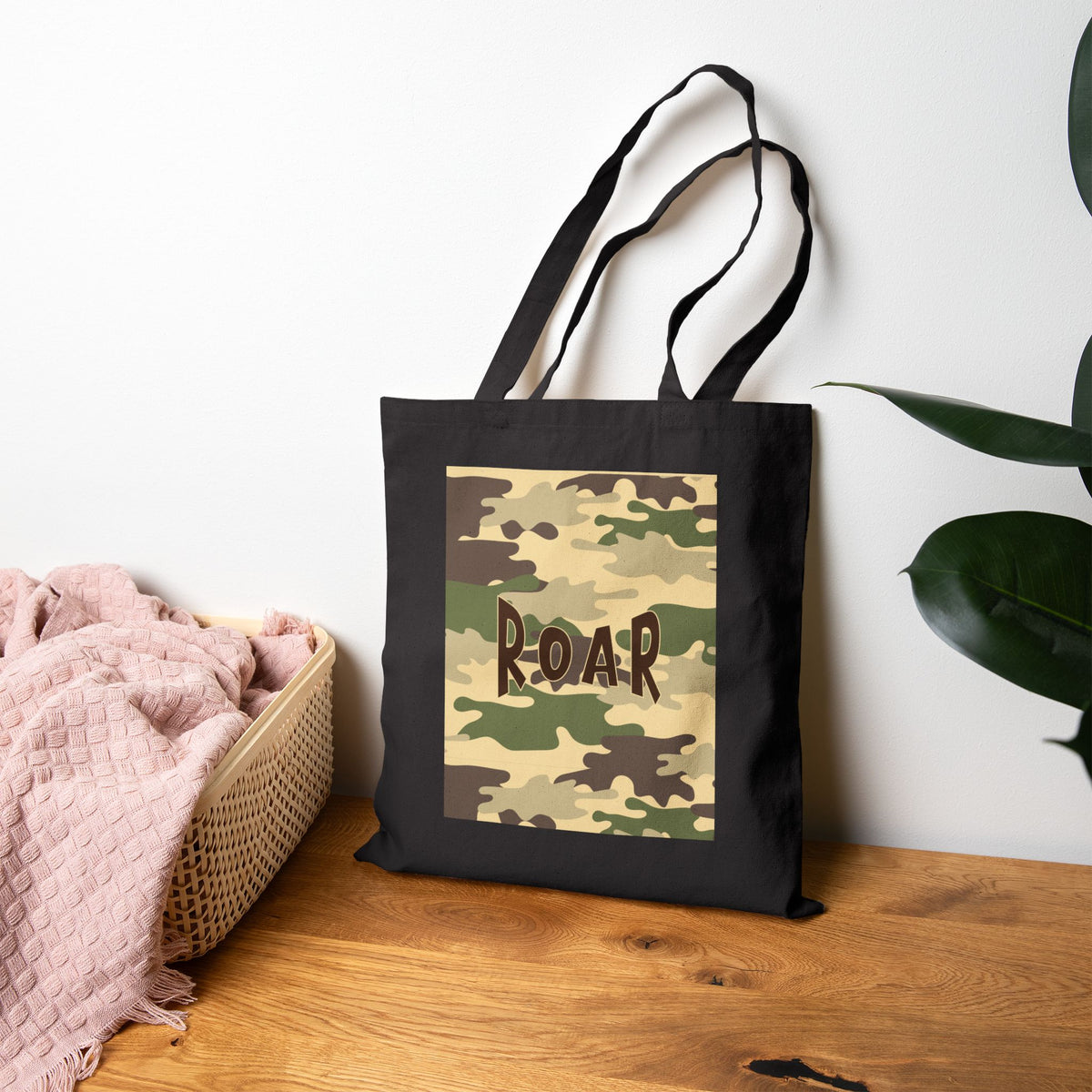 A black canvas tote bag with a camouflage print panel on the front featuring the word 'ROAR' in brown letters, displayed on a wooden table next to a pink blanket and a green plant against a white wall.