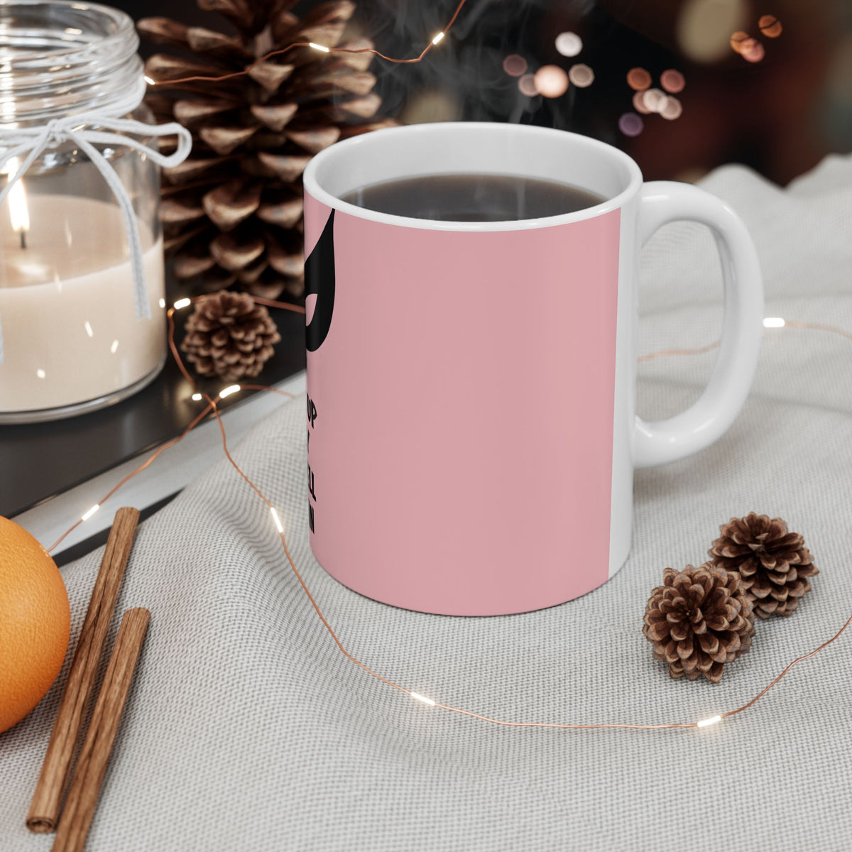A pink ceramic mug with a white handle and interior, filled with a dark beverage and placed on a light fabric surface. The mug has a black graphic on the side.