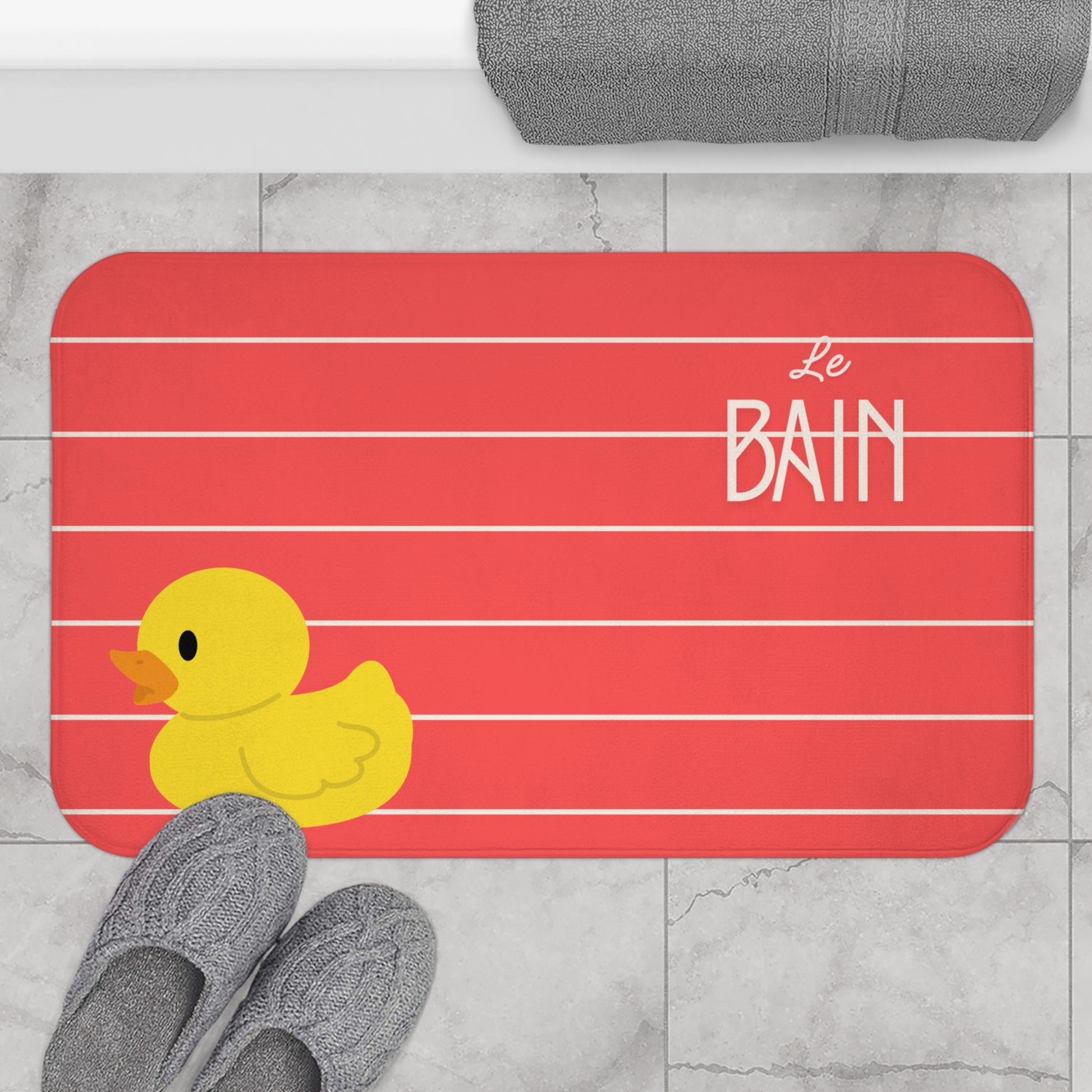 Bath mat with a yellow rubber duck and 'Le Bain' text on a red background, placed on a tiled floor.