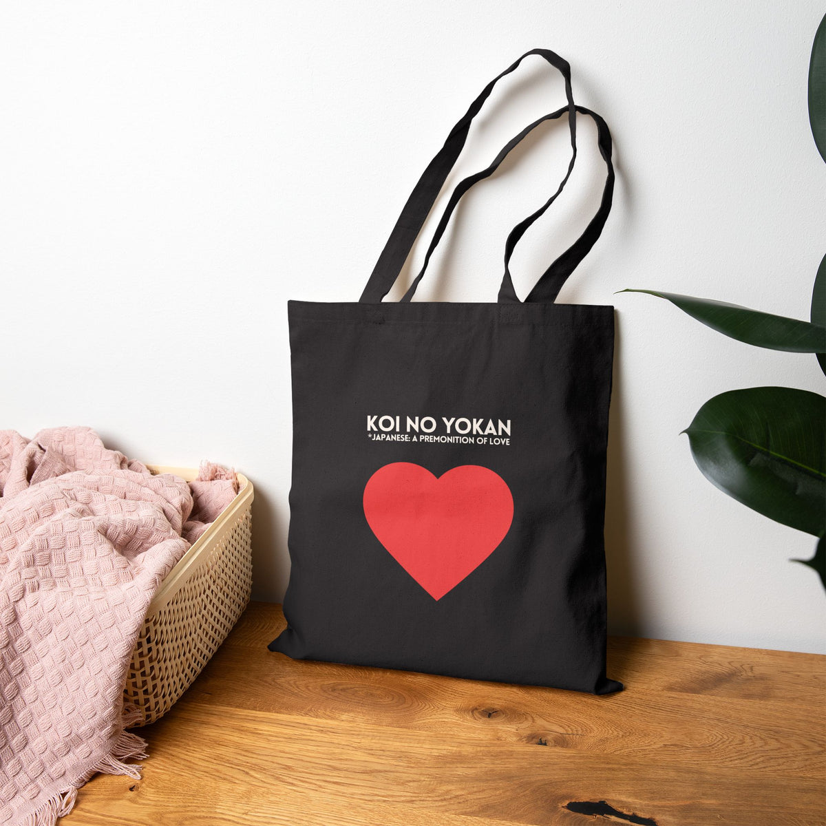 A black canvas tote bag with long handles, featuring the white text 'KOI NO YOKAN' and a large red heart on the front. The bag is displayed on a wooden table against a white wall, styled with a pink blanket in a basket on the left and a green plant on the right.