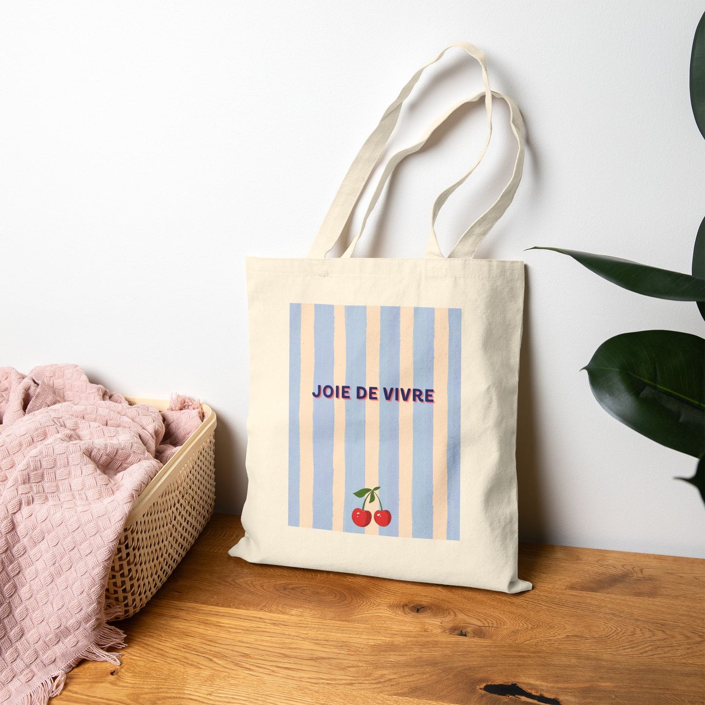 Beige tote bag with a nature-themed design on a wooden surface with a white wall and pink blanket in the background.