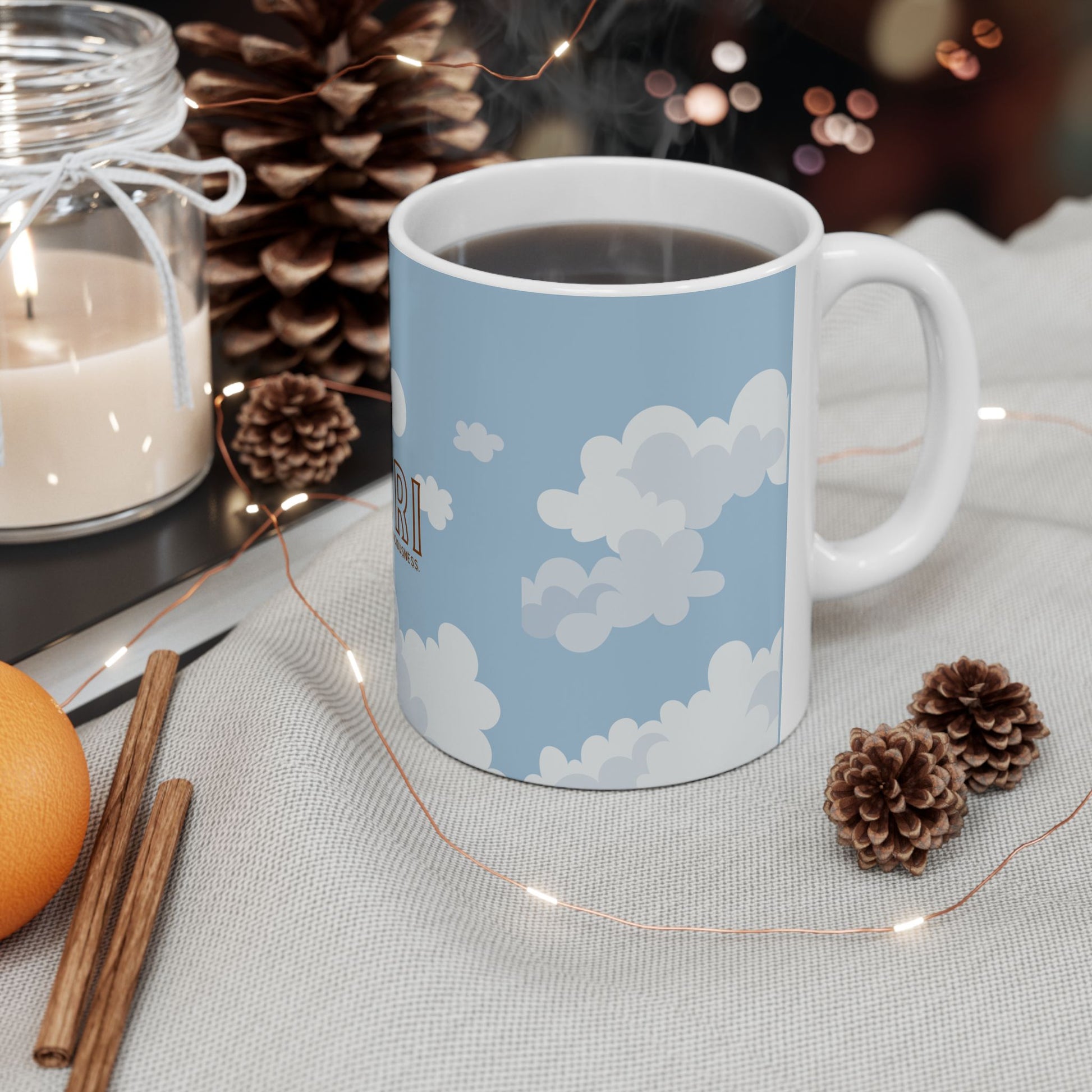 A white ceramic mug with a light blue sky and white cloud print, filled with a hot beverage and set on a textured light gray cloth with pinecones, fairy lights, a candle, cinnamon sticks, and an orange.