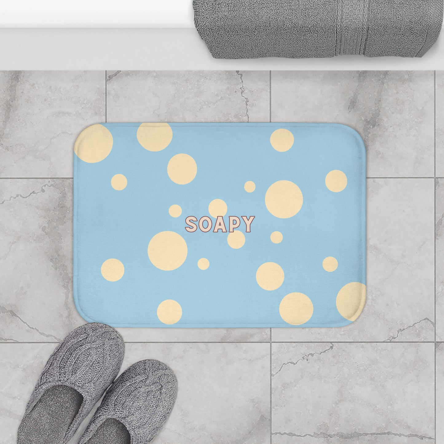 A rectangular blue bath mat with yellow polka dots and the word 'SOAPY' in the center, shown on a gray marble tile floor with gray slippers and a folded gray towel nearby.