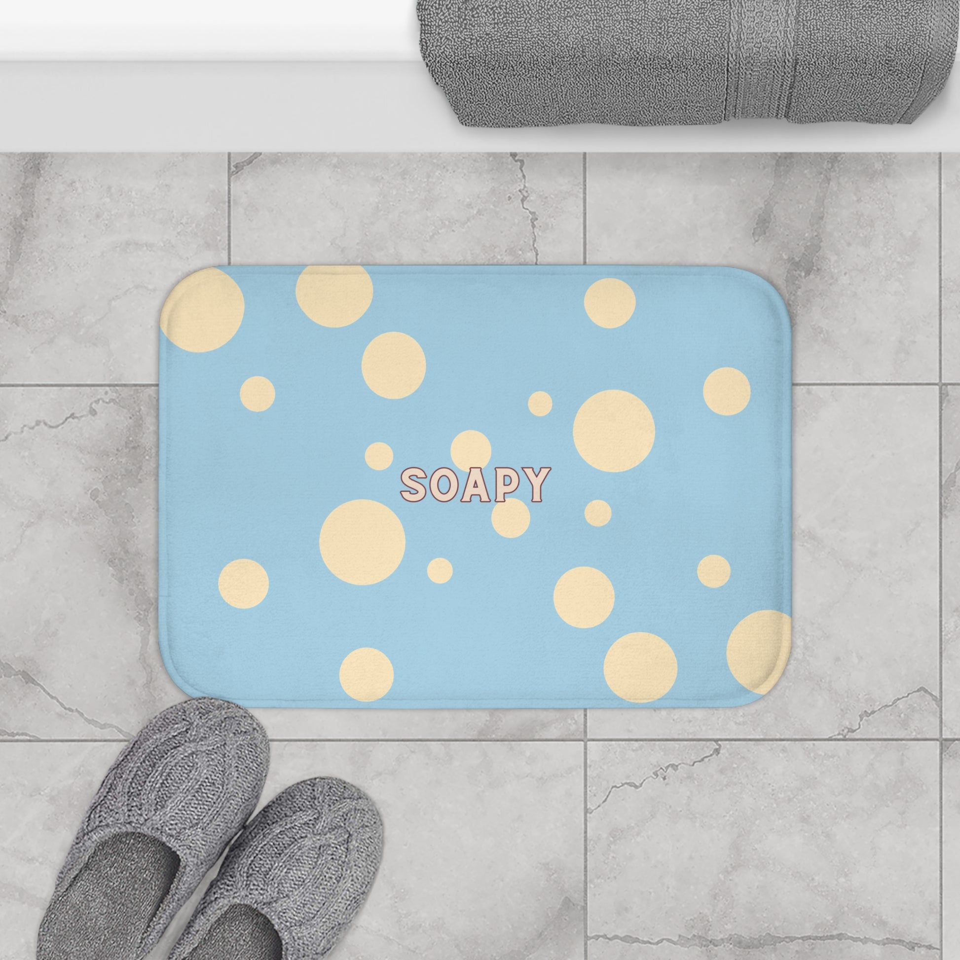 A rectangular blue bath mat with yellow polka dots and the word 'SOAPY' in the center, shown on a gray marble tile floor with gray slippers and a folded gray towel nearby.
