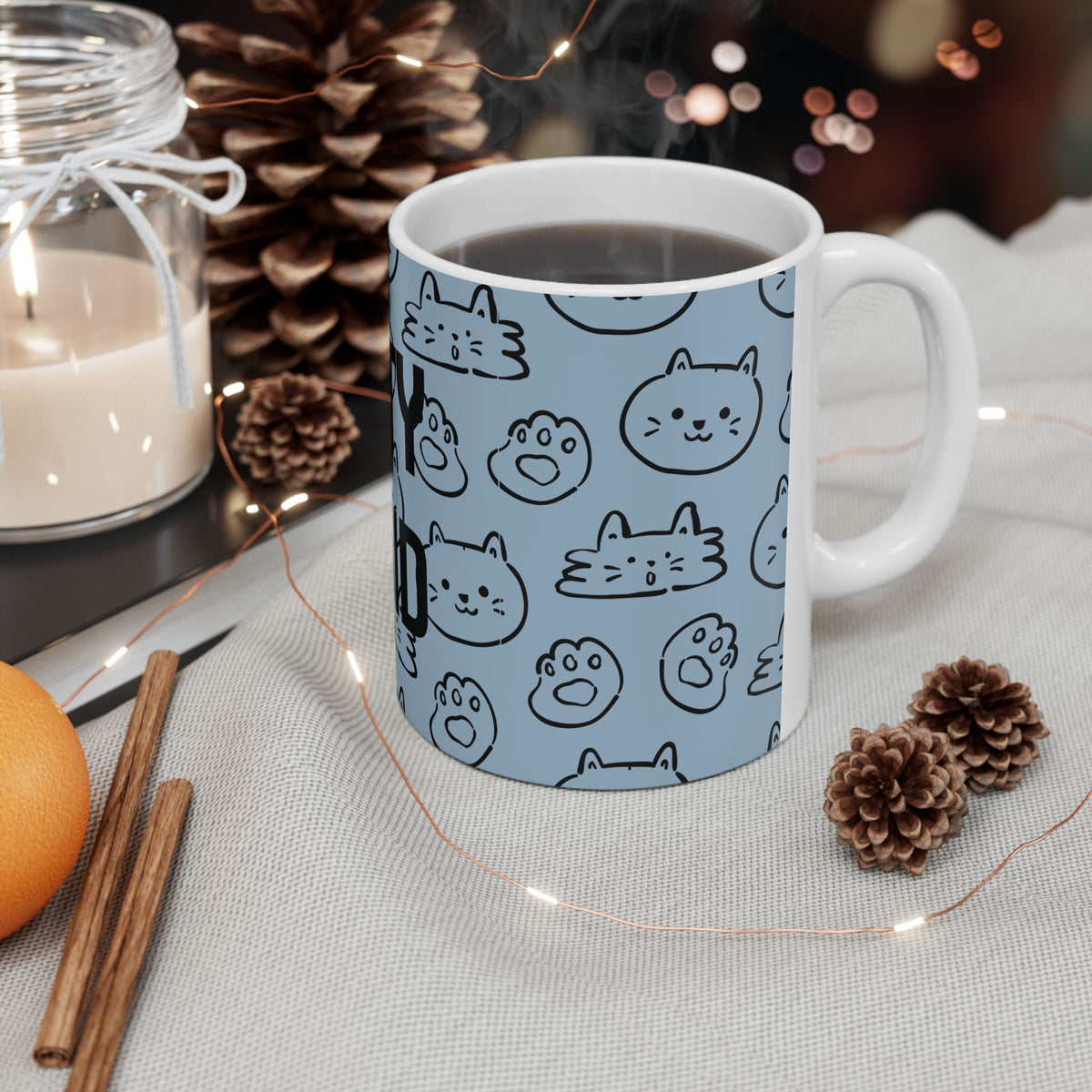 A white ceramic mug filled with a dark beverage, decorated with a light blue wraparound print of black cat face and paw prints. The mug is styled on a light fabric surface with warm fairy lights, pinecones, cinnamon sticks, and a candle in the background.