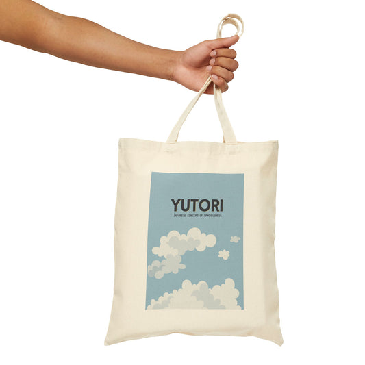 An off-white canvas tote bag with a light blue graphic print featuring the text 'YUTORI' and clouds, held by a person's hand against a white background.