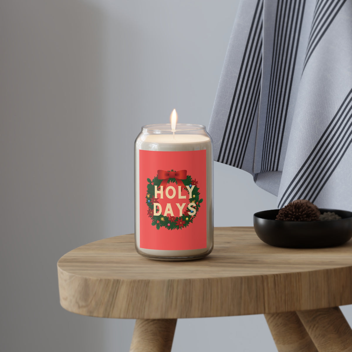 A lit candle in a clear glass jar sits on a wooden stool beside a black bowl, with a gray wall and striped fabric in the background. The candle label reads “HOLY DAYS” in cream text inside a green wreath with red accents.