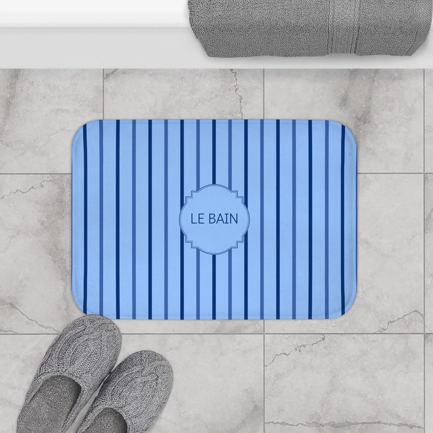 A rectangular blue bath mat with vertical stripes and a central medallion reading 'LE BAIN', shown on a light marble bathroom floor with gray towels and slippers nearby.