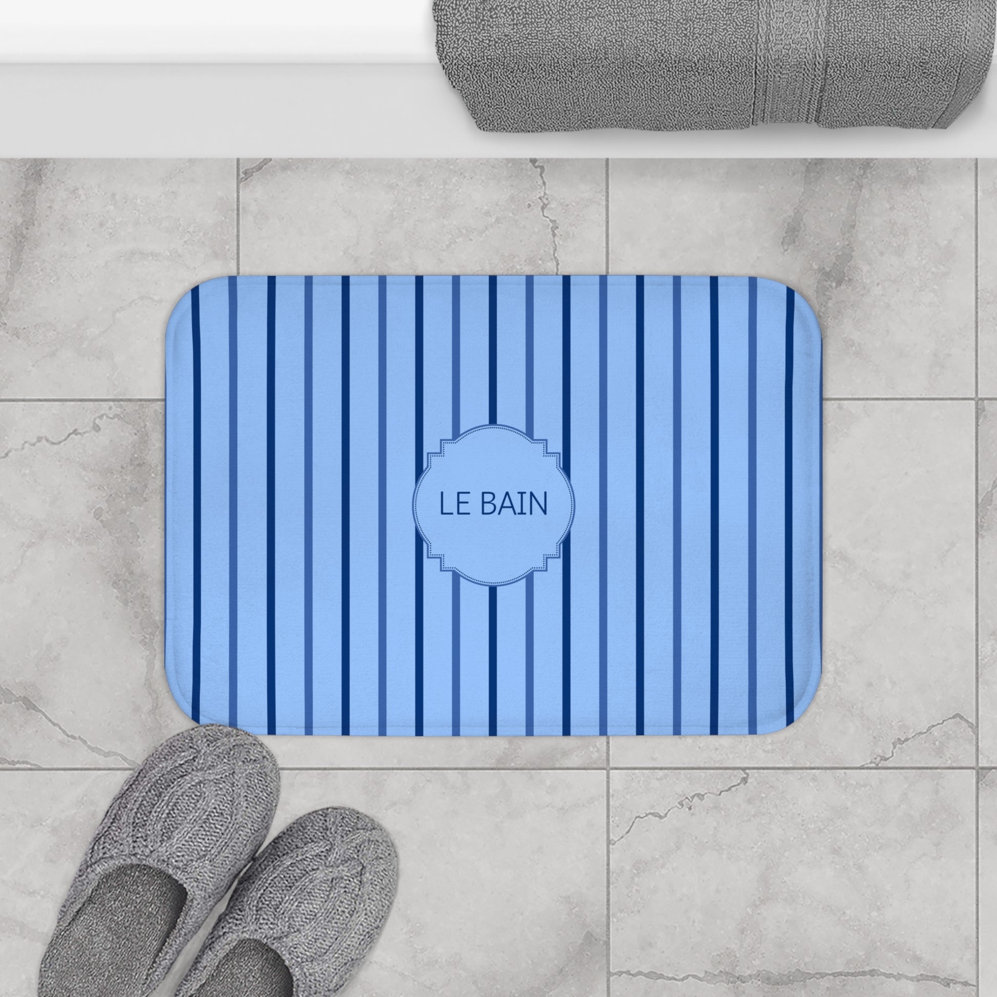 A rectangular blue bath mat with vertical stripes and a central medallion reading 'LE BAIN', shown on a light marble bathroom floor with gray towels and slippers nearby.