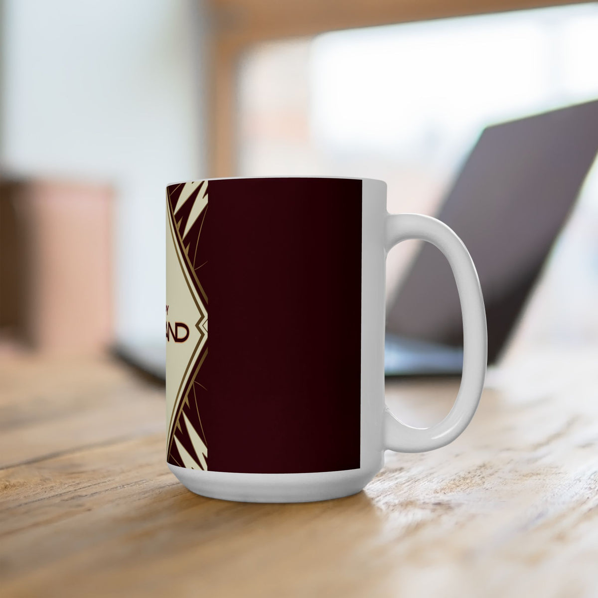A white ceramic mug with a dark maroon wraparound print and a cream geometric side design, sitting on a wooden table with a blurred laptop in the background.