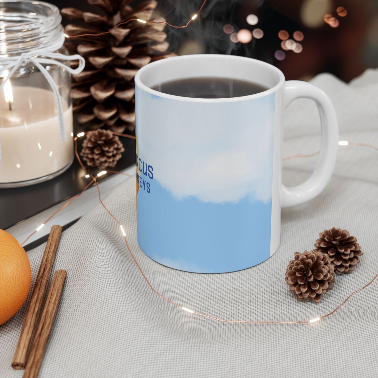 A white ceramic mug filled with a dark beverage, decorated with a light blue and white landscape print, styled on a light fabric surface with pinecones, fairy lights, a candle, cinnamon sticks, and an orange.