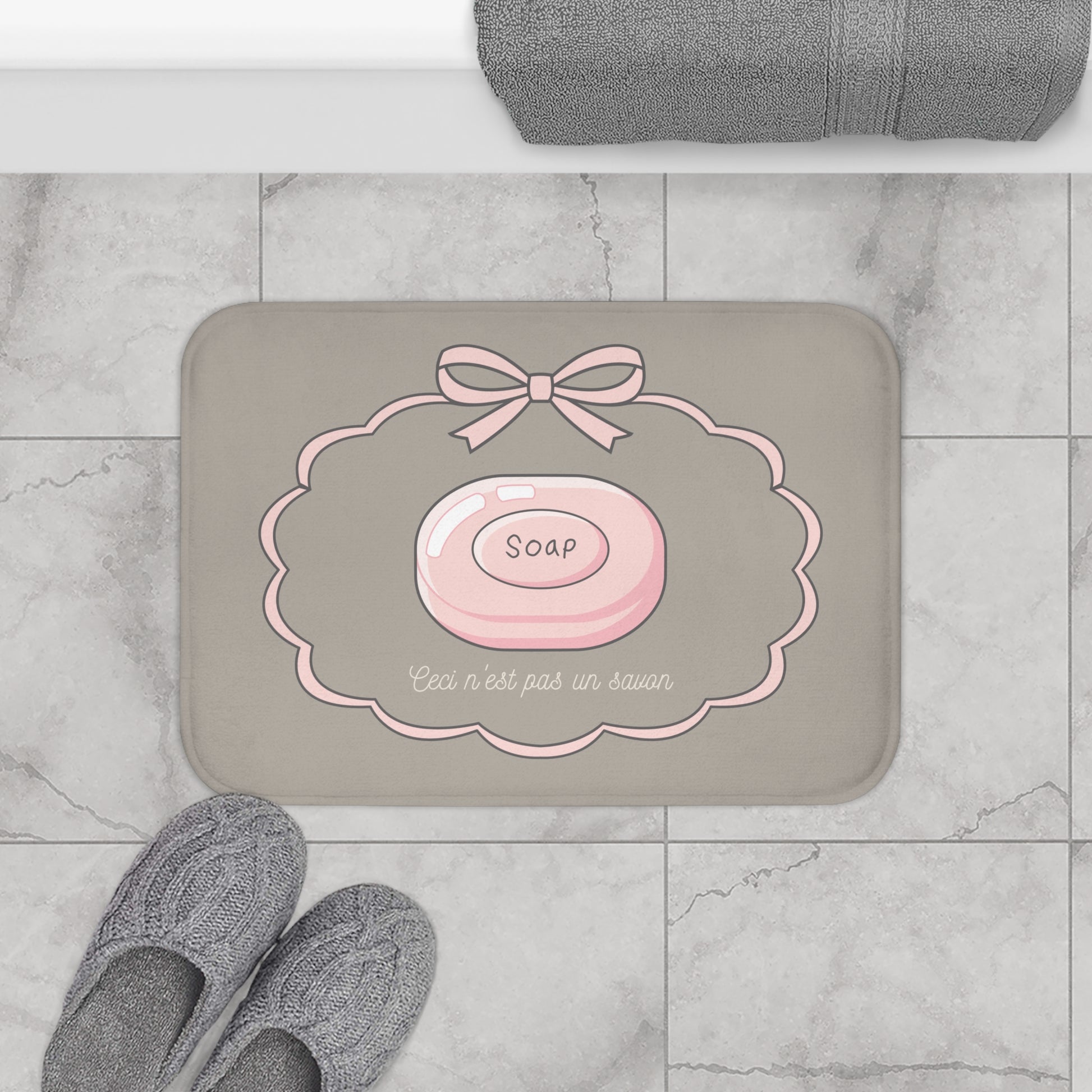Gray bath mat with pink soap design on a marble floor with slippers and a towel.