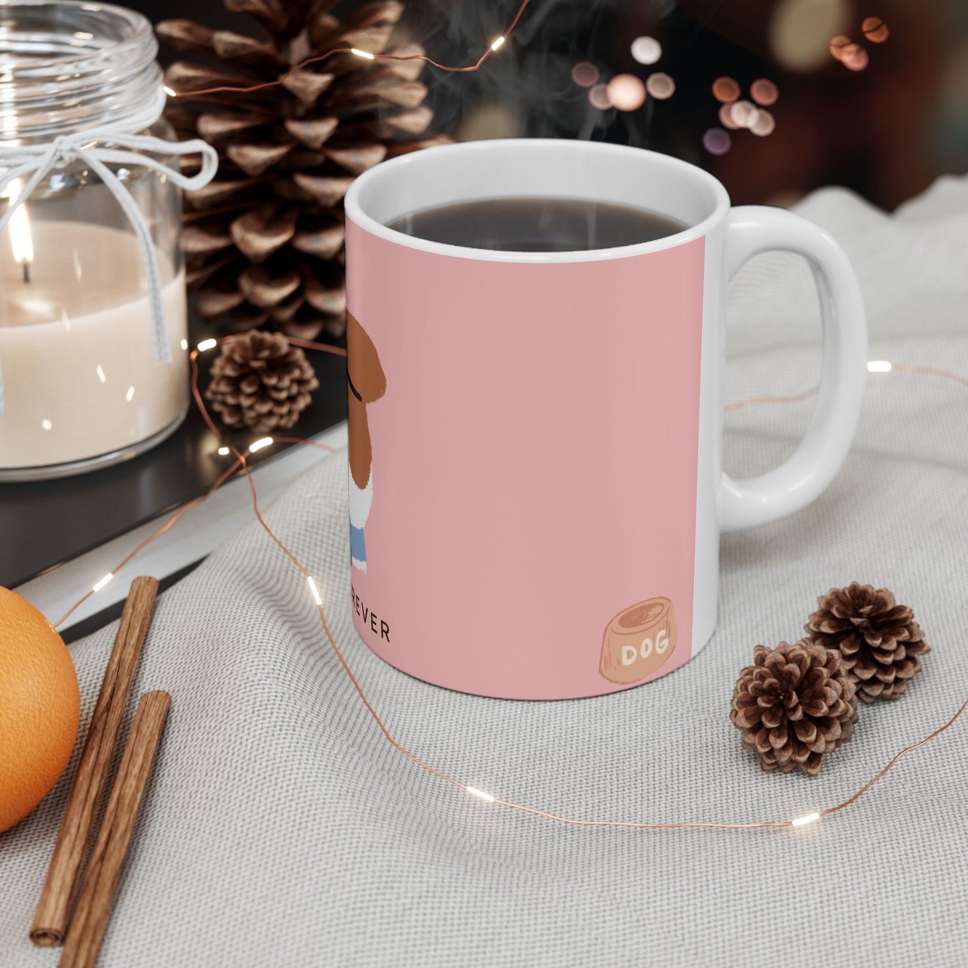 A pink ceramic mug with a white handle and base, filled with a hot beverage and surrounded by cozy holiday props including pinecones, fairy lights, a candle, cinnamon sticks, and an orange on a light fabric surface.