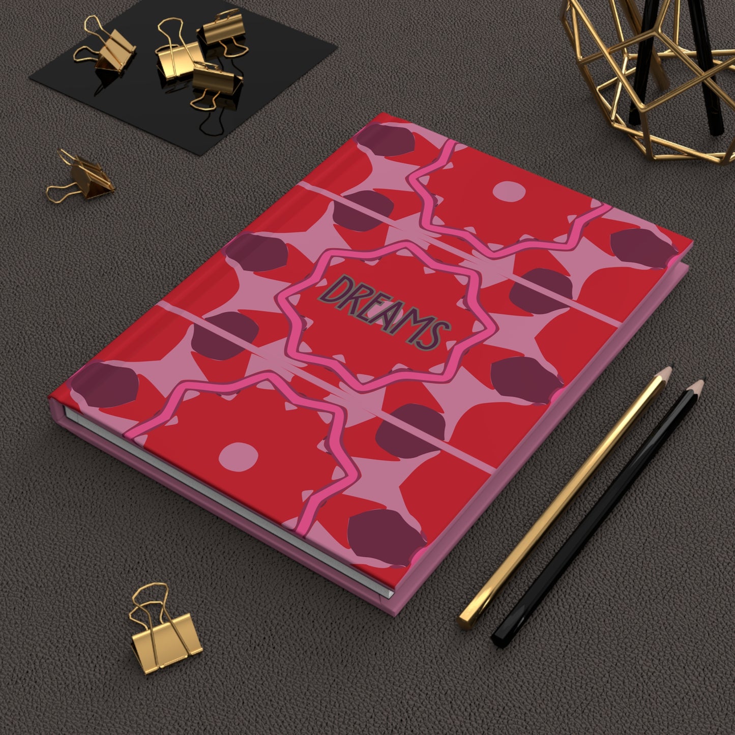 Notebook with red and pink abstract design and 'DREAMS' text on a dark surface with office supplies.