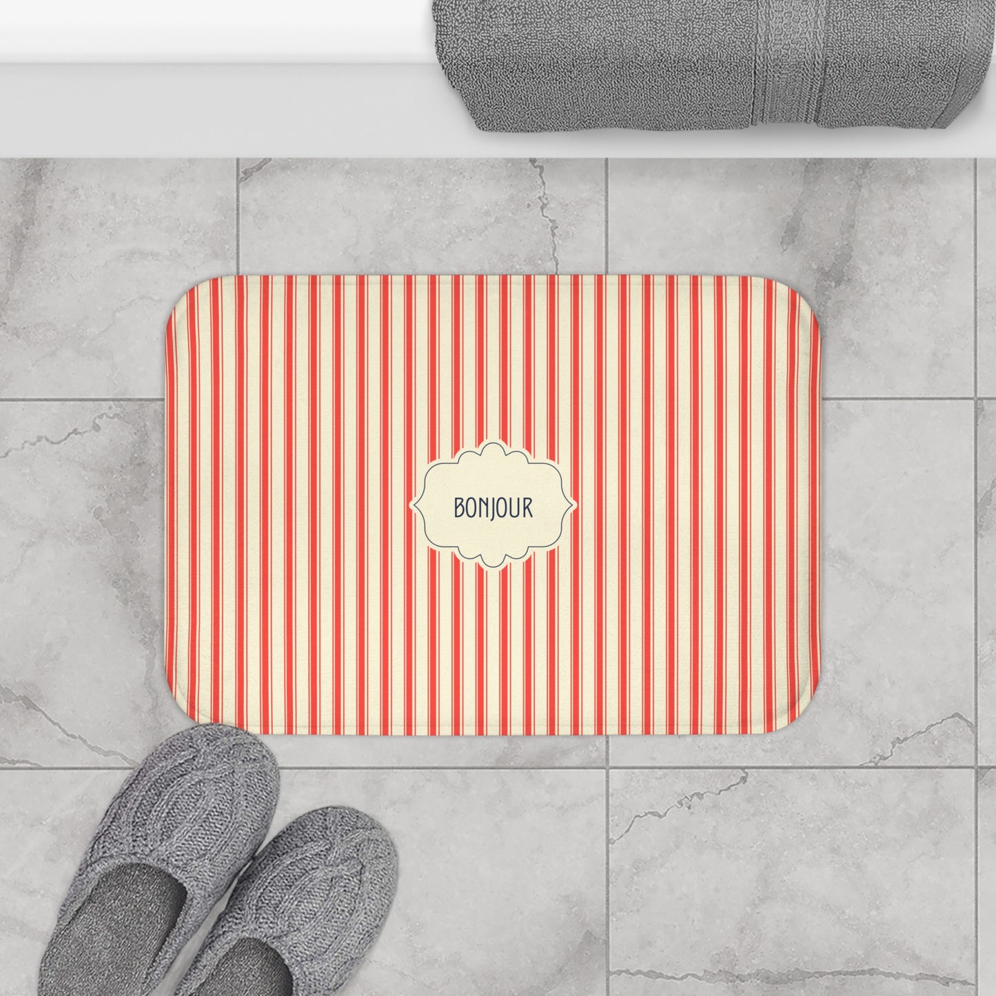 A rectangular bath mat with red and white vertical stripes and a cream label in the center reading 'BONJOUR', shown on a gray marble tile floor with gray slippers and a folded gray towel nearby.