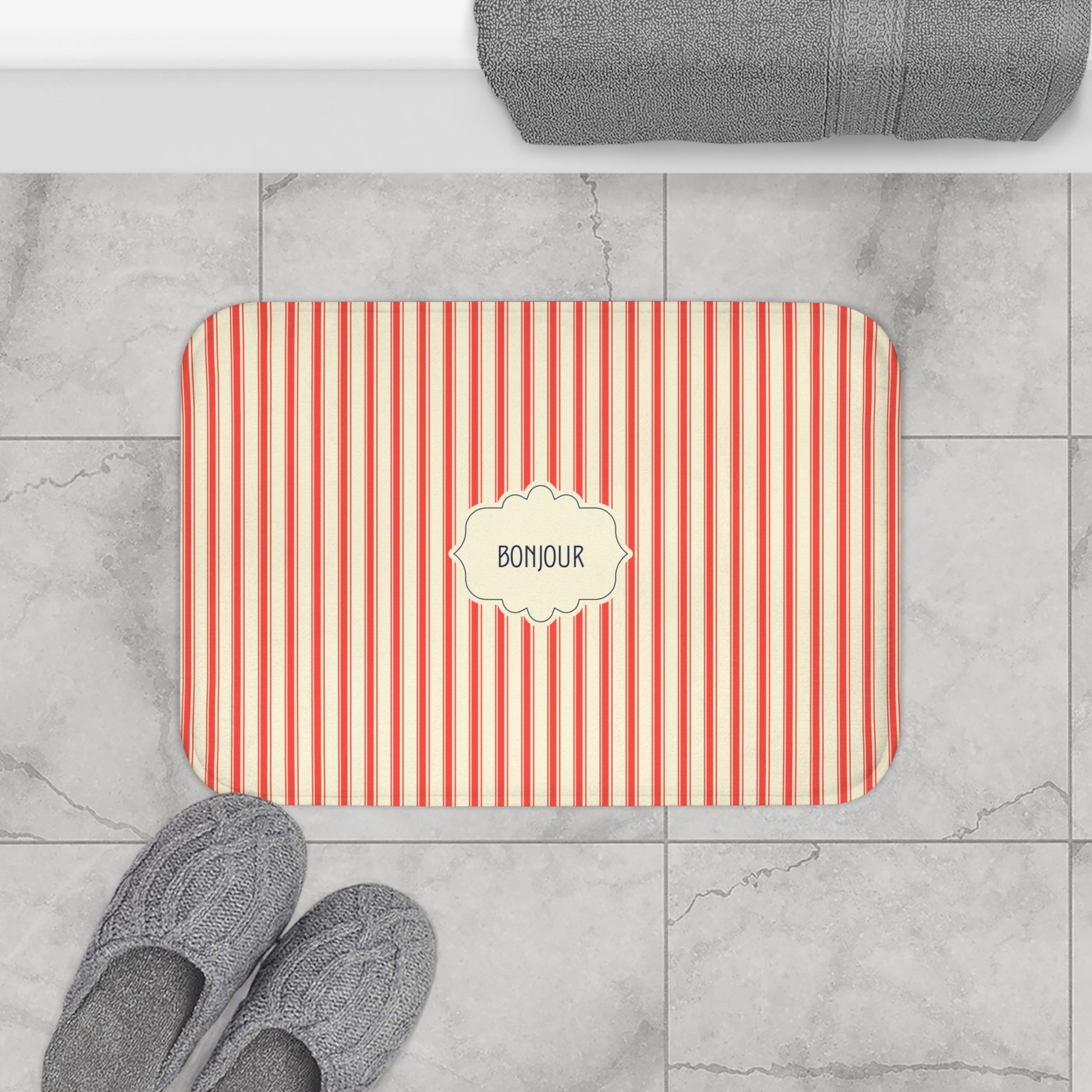 A rectangular bath mat with red and white vertical stripes and a cream label in the center reading 'BONJOUR', shown on a gray marble tile floor with gray slippers and a folded gray towel nearby.