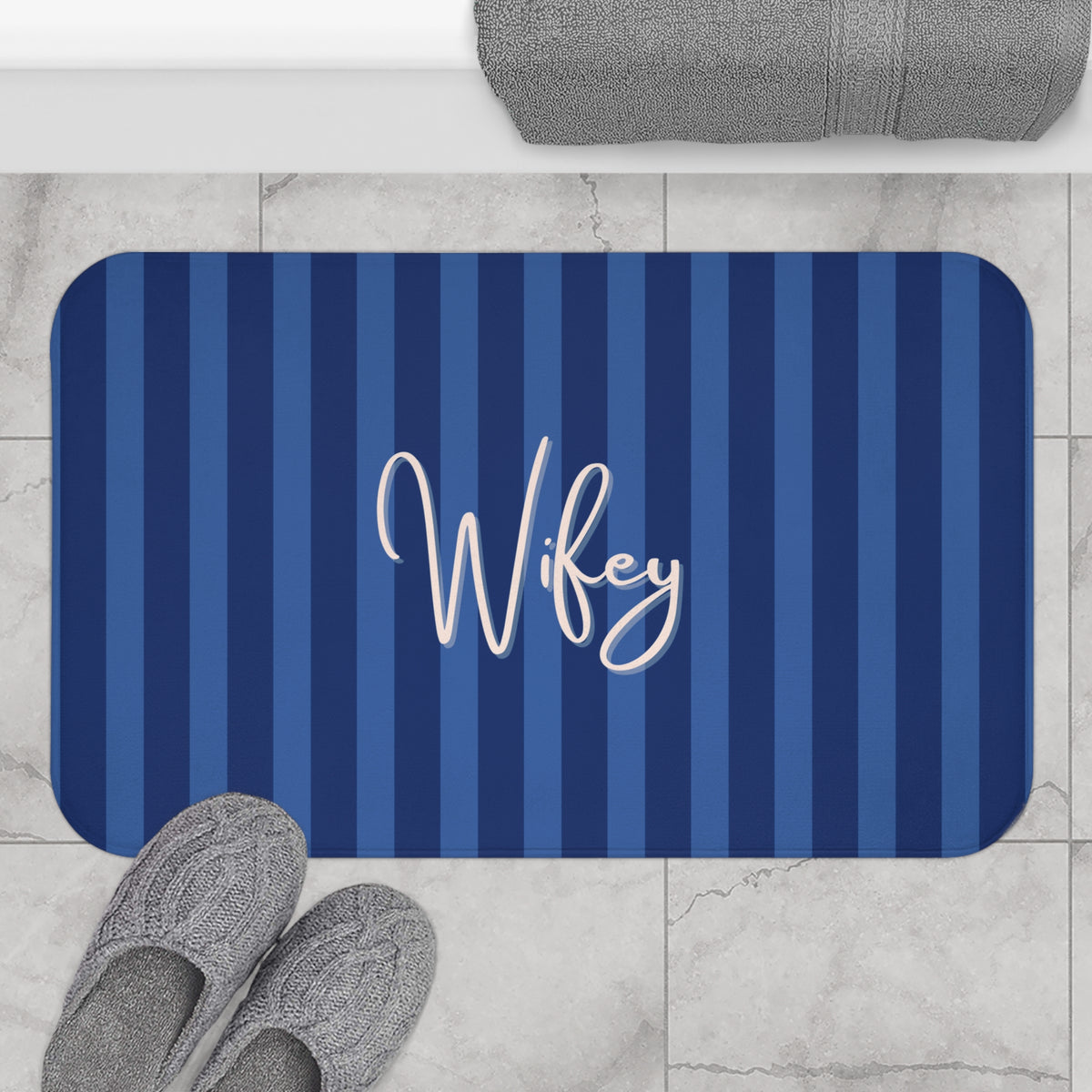 A rectangular bath mat with rounded corners, featuring vertical navy and medium blue stripes and the word 'Wifey' in white script across the center. The mat is shown on a light marble tile floor with gray slippers and a folded gray towel nearby.