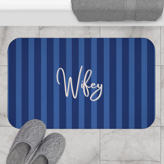 A rectangular bath mat with rounded corners, featuring vertical navy and medium blue stripes and the word 'Wifey' in white script across the center. The mat is shown on a light marble tile floor with gray slippers and a folded gray towel nearby.