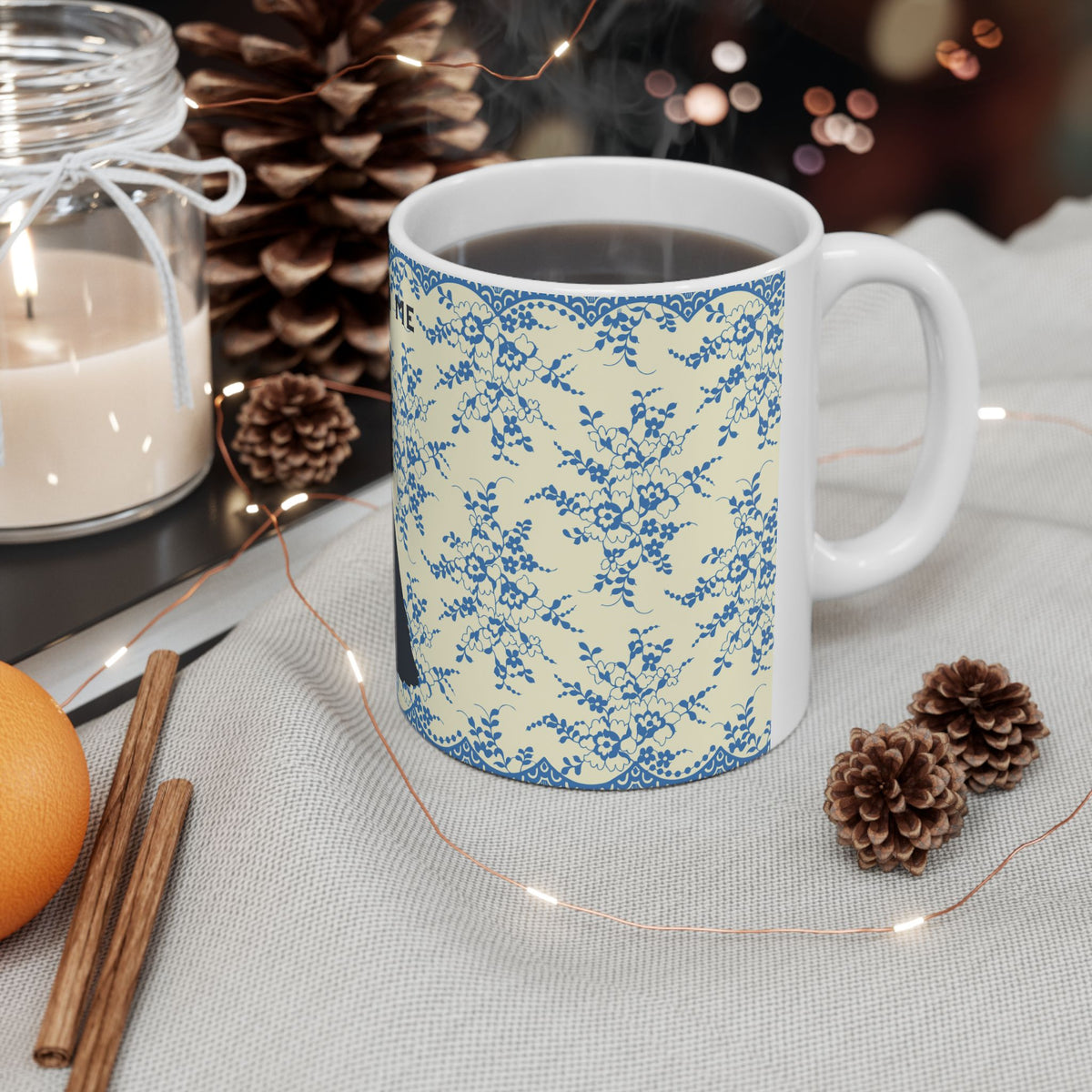 White mug with hot chocolate, surrounded by decorative elements like a candle, cinnamon sticks, and pine cones on a table.