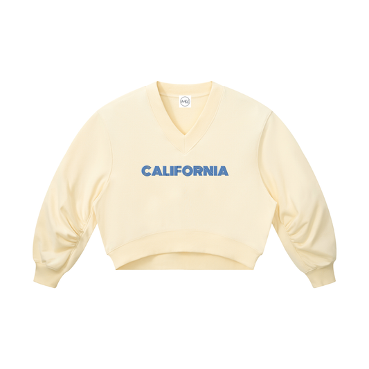 A pale cream-colored cropped V-neck sweatshirt with 'CALIFORNIA' printed in blue across the chest, displayed on a solid white background.